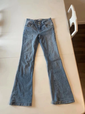 Ljusblå bootcut jeans - Säljer ett par ljusblå bootcut jeans med klassisk femficksdesign och knappstängning. Jeansen har en snygg utsvängd passform nedtill och är tillverkade i denim med en mjuk känsla. Perfekta för dig som gillar retrovibbar och vill ha ett par bekväma jeans.Ljusa denimjeans i storlek 158 cm 💙 Low-rise/låg till medelhög midja och detaljerade fickor på sidorna. Passar XXS–XS beroende på önskad passform. Perfekta för Y2K, 2000s, low-rise- midrise och vintage outfits 🩵  y2k • 2000s • low rise • l