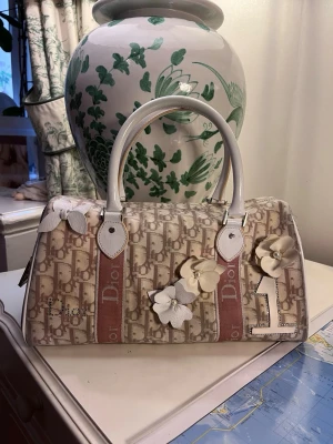 Dior handväska med blommor - Säljer en handväska från Dior den heter Mini Boston Trotter med det klassiska Dior monogrammönstert och ett axelband, som jag tillverkat själv. Väskan har vita handtag, detaljer i vitt skinn, så  som dekorativa blommor och rosett i vitt skinn och med strass och namnet Dior framtill. Dior hänglås sitter på också men det kan jag skicka separata bilder på om du vill? Dustbag finns i original. Den är till dig som vill ha vara Miss Dior hela dagen.