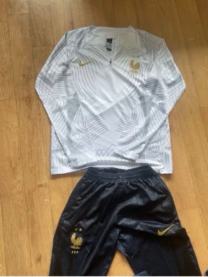 Frankrike bara byxor - Snyggt träningsset från Nike med Frankrikes landslagslogga. Tröjan är vit med grått grafiskt mönster, halv dragkedja och lång ärm. Byxorna är svarta med gulddetaljer och resår i midjan. Perfekt för fotbollsträning eller chill.