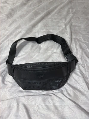 Svart shoppingväska från Giorgio Armani - Snygg svart shoppingväska från Giorgio Armani med justerbart band och dragkedja. Väskan har ett diskret mönster med logotypen tryckt över hela ytan och på axelremmen. Tillverkad i slitstarkt material som ger en modern och stilren look.