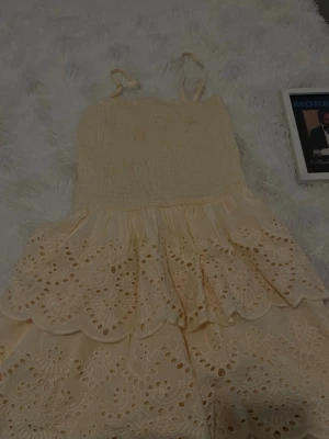 Ljusgul topp med broderade volanger - Söt ljusgul topp med smock upptill och tunna axelband. Den har flera lager med broderade volanger och hålmönster som ger en romantisk känsla. Perfekt för varma dagar och somriga tillfällen.