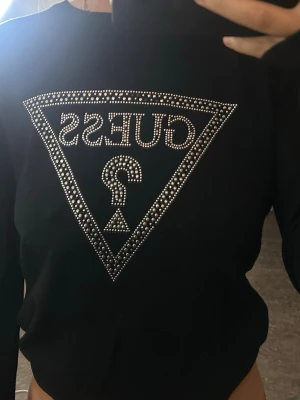 Svart långärmad topp från Guess - Snygg svart långärmad topp från Guess med det ikoniska trekantsloggan framtill, dekorerad med silvriga nitar som ger en lyxig känsla. Tillverkad i mjukt material och har en klassisk passform. Perfekt för dig som vill sticka ut med en trendig och stilren look.