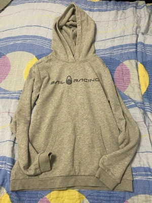 Grå hoodie från Sail Racing - Snygg grå hoodie från Sail Racing med svart logga tryckt på bröstet. Klassisk känguruficka framtill och ribbade muddar vid ärmar och nederkant. Tillverkad i mjukt sweatshirtmaterial och har en stor huva för extra komfort.
