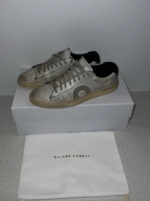 Oliver cabell  - Snygga vita sneakers från Oliver Cabell med grå cirkeldetalj på sidan och svart hälparti. Skorna har en klassisk låg siluett, beige sula och är tillverkade i läder. Perfekta för dig som gillar stilrena men ändå unika sneakers.