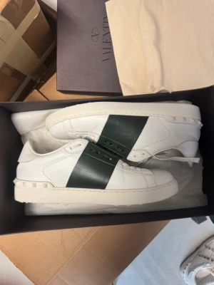 Valentino Opens  - Snygga vita sneakers med bred grön kahki färg rem över snörningen och nitar längs hälen. Skorna har en klassisk låg profil och är tillverkade i slätt läder. Perfekta för dig som vill ha en stilren men ändå unik look. Endast skor ingår! Hör av dig vid minsta fråga🙌