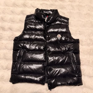 Moncler Väst - Snygg svart dunväst från Moncler med glansig finish och klassisk logga på bröstet. Västen har hög krage, två dragkedjeförsedda fickor och quiltad design. Perfekt för lager-på-lager och streetstyle. Materialet är dunfyllning och yttertyget är blankt.