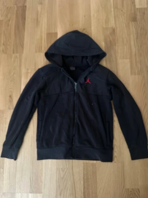 Nike Jordan kofta - Säljer en svart hoodie med dragkedja och huva. Hoodien har en röd logga på bröstet och två fickor framtill. Tillverkad i mjukt material och har långa ärmar samt ribbade muddar. Perfekt för en avslappnad och sportig stil. Det finns vita prickor den är lite smutsigt men går bra att tvätta till helt ny! 6/10 men om man tvättar blir det 8/10