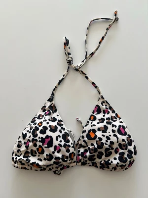 Leopardmönstrad bikiniöverdel med knyt - Snygg trekantsbikini med leopardmönster i svart, brun, orange och rosa på vit botten. Överdelen har knyt både i nacken och ryggen för justerbar passform. Perfekt för sommaren och strandhäng! Aldrig använd!