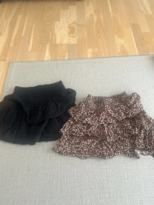 Två korta volangkjolar - Säljer två snygga kortkjolar, en svart med volanger från Gina Tricot i stl 146/152 och en brun med vitt blommönster och volangdetaljer från Lindex i stl 134/140 Perfekta för dig som gillar trendiga och feminina plagg. Båda har resår i midjan för skön passform.