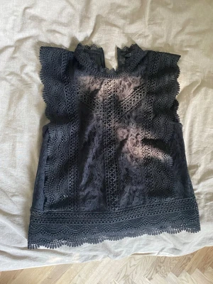 Svart spetsblus med volangdetaljer - Supersnygg svart blus i spets med volangdetaljer längs ärmhål och nederkant. Blusen är ärmlös och har vackra broderade mönster som ger en romantisk känsla. Perfekt att styla med jeans eller kjol för en trendig look.
