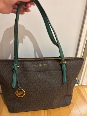 Brun Michael Kors handväska - Snygg brun handväska från Michael Kors med klassiskt MK-mönster och gröna axelband. Väskan har guldfärgade detaljer och dragkedja upptill. Perfekt storlek för att få plats med allt du behöver. Materialet är tåligt konstläder.