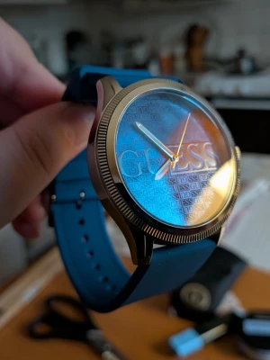 Blå klocka från Guess med silikonband - Snygg blå klocka från Guess med silikonband och silverfärgat boett. Urtavlan har Guess-loggan i stort och ett upprepat mönster med mindre logotyper. Klockan har en modern och stilren design som sticker ut.