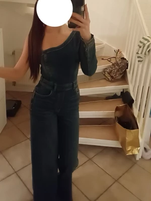 Mörkblå one shoulder denim jumpsuit - Säljer en snygg mörkblå denim jumpsuit med one shoulder-design och långa vida ben. Jumpsuiten har en ärm och markerad midja, samt klassiska jeanssömmar och fickor fram. Perfekt för dig som vill sticka ut med en unik look.