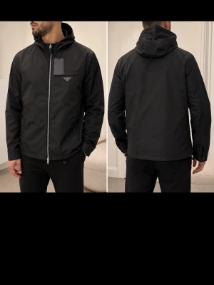 Svart vindjacka med huva - Snygg svart vindjacka med huva och dragkedja framtill. Jackan har en minimalistisk design, diskret bröstficka med logga och två sidofickor. Tillverkad i ett lätt, vattenavvisande material som passar perfekt för blåsiga dagar.