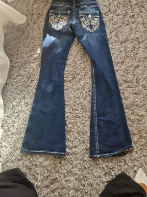 Blå bootcut jeans med broderade fickor - Säljer ett par mörkblå bootcut jeans med snygga slitningar och kontrastsömmar. Baksidan har två broderade fickor med dekorativa detaljer och knappar. Jeansen har låg midja och klassisk femficksmodell. Perfekta för en trendig och avslappnad stil.