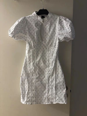 Vit prickig kortklänning Gina Tricot - Supersöt vit kortklänning från Gina Tricot med svarta prickar, puffärmar och en snygg cut-out framtill. Klänningen har en figurnära passform och hög hals. Perfekt för dig som vill sticka ut lite extra!