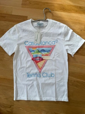 Vit Casablanca Tennis Club t-shirt - Säljer en vit t-shirt från Casablanca med färgglatt grafiskt tryck och texten 'Tennis Club' på framsidan. T-shirten har rund hals och korta ärmar. Perfekt för dig som gillar sportig och trendig stil.