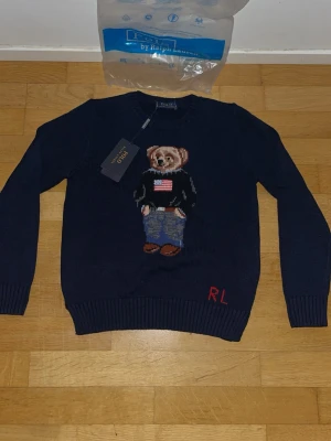 Mörkblå stickad tröja med björn - Mörkblå stickad tröja från Ralph Lauren med ikonisk björn på bröstet, klädd i jeans och svart tröja med amerikansk flagga. RL-broderi i rött nedtill. Rund halsringning och ribbade muddar. Perfekt för dig som gillar klassisk preppy stil.