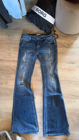 Blå bootcut jeans från meet me there  - Säljer ett par blå bootcut jeans med slitningar och hål på benen. Jeansen har klassisk femficksdesign, låg midja och dragkedja med knapp. Perfekta för en avslappnad och trendig look. Har aldrig använt då de är lite för små 