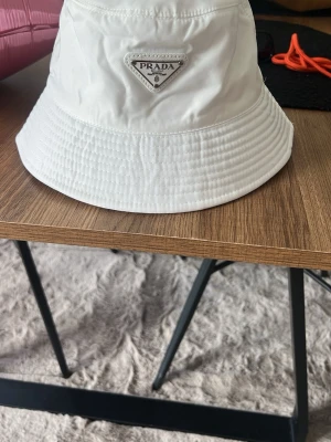 Vit bucket hat från Prada - Säljer en stilren vit bucket hat från Prada med det klassiska trekantiga logotyp-emblemet framtill. Hatten är tillverkad i ett glansigt, lätt material som ger en modern vibe och passar perfekt till streetwear eller sommaroutfits.