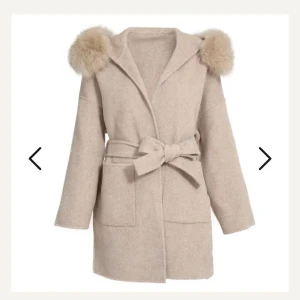 Beige kappa med fuskpälsdetaljer - Trendig beige kappa med stora fickor, knytskärp i midjan och fluffig fuskpäls på luvan . Rak passform och mjukt material som ger en lyxig känsla. Perfekt för dig som vill ha en stilren men ändå unik look. Perfekt nu till hösten!!