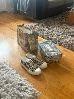 Dior sneakers med monogrammönster - Snygga Dior sneakers i vitt och svart med klassiskt monogrammönster och vita snören. Skorna har låg profil och rund tå, samt tydlig logga på plösen och sidan. Perfekta för dig som vill ha en exklusiv och trendig look.