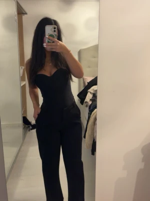 Svart byxdress med sweetheart-ringning - Säljer en svart byxdress med hjärtformad urringning. Byxdressen har vida ben och är axelbandslös, vilket ger en clean och stilren look. Perfekt för dig som vill ha något enkelt men ändå snyggt. Materialet ser ut att vara mjukt och följsamt.