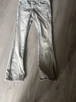 Grå bootcut jeans med låg midja - Säljer ett par grå bootcut jeans med låg midja och justerbar midja. Jeansen har dubbla silverknappar framtill och klassiska fickor. Materialet är en blandning av bomull, polyester och elastan för extra stretch och komfort.
