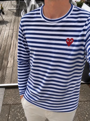 Comme des Garçons PLAY Striped Long Sleeve  - Skapa en avslappnad och stilren look med denna ikoniska långärmade tröja från Comme des Garçons PLAY. Plagget har ett klassiskt blåvitt Breton-randigt mönster i mjuk bomull av hög kvalitet och pryds av den ikoniska, broderade röda hjärtlogotypen på bröstet, vilket ger en omedelbar designer-känsla. En riktig garderobsklassiker som tillför en subtil retrokänsla till vilken outfit som helst – kombinera med dina favoritbyxor eller jeans. 