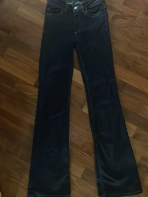 Mörkblå bootcut jeans med kontrastsöm - Säljer ett par mörkblå bootcut jeans med vita kontrastsömmar och en unik broderad detalj på bakfickan. Klassisk femficksmodell i denim med hög midja och utsvängda ben. Perfekta för dig som gillar retrovibbar och vill sticka ut lite extra. Nästan helt nya men använd ett par gånger. Nypris 899