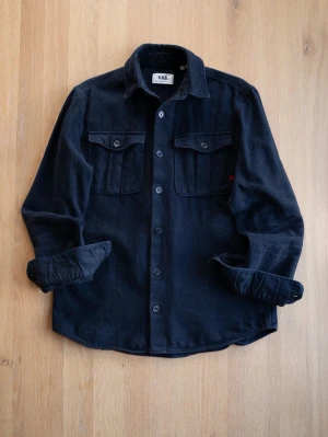 Black Heavyweight Overshirt, size S - Stilren svart overshirt från RDD med två bröstfickor med lock och knappar. Skjortan har klassisk krage, raka ärmar och knäppning framtill. Tillverkad i ett mjukt, tjockare tyg som passar perfekt som lager på lager.