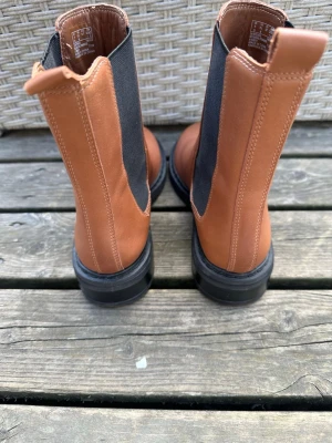 Bruna Chelsea boots från Clarks - Snygga Chelsea boots i brunt läder från Clarks med svarta elastiska paneler på sidorna och grov sula. Klassisk siluett med dragflikar både fram och bak för enkel påtagning. Perfekta för dig som vill ha en stilren och tidlös look.