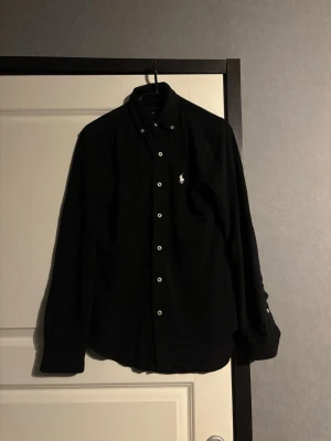 Svart skjorta med knappar - Stilren svart skjorta med långa ärmar och vita knappar framtill. Klassisk krage och diskret broderad logga på bröstet. Perfekt för dig som gillar en enkel men snygg look.