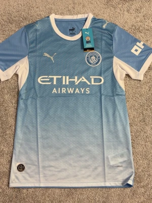 Manchester City matchtröja Puma - Snygg ljusblå Manchester City matchtröja från Puma med vita detaljer och klubbmärke på bröstet. Tröjan har Etihad Airways-tryck på framsidan och är tillverkad i ett lätt, funktionsmaterial (Drycell) som andas. Perfekt för fotbollsträning eller att bära till vardags.