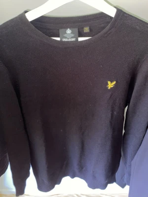 Svart stickad tröja från Lyle & Scott - Klassisk svart stickad tröja från Lyle & Scott med rund halsringning och den ikoniska gula örnloggan broderad på bröstet. Tröjan har långa ärmar och ribbade muddar vid ärmslut och nederkant. Perfekt basplagg till garderoben.