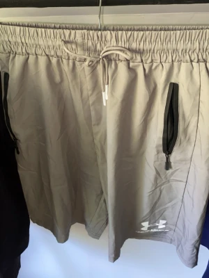 Beige shorts från Under Armour - Snygga beige shorts från Under Armour med elastisk midja och snörning. Två svarta dragkedjefickor fram och en bakficka. Perfekta för sommarens alla aktiviteter och tillverkade i ett lätt syntetmaterial.