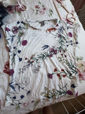 Vit t-shirt med blommigt mönster - Säljer en vit t-shirt med färgglatt blommigt mönster i lila, rött, blått, grönt och gult. T-shirten har korta ärmar och är gjord i mjuk bomull. Perfekt för dig som gillar en fräsch och somrig stil med mycket färg.