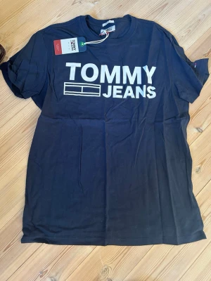 Svart t-shirt från Tommy Jeans - Snygg svart t-shirt från Tommy Jeans med stort vitt tryck på bröstet. Klassisk passform och rund halsringning. Perfekt till jeans eller shorts för en clean och enkel look. Helt oanvänd. 