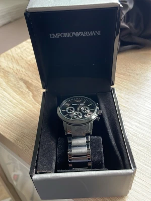 Emporio Armani svart klocka - Stilren svart klocka från Emporio Armani med metallarmband och flera små urtavlor på svart urtavla. Klockan har en klassisk rund form och detaljer i silver. Perfekt accessoar för dig som gillar en elegant och modern look.
