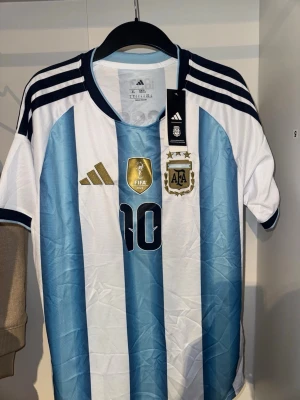 Argentina Messi matchtröja Adidas - Säljer en officiell Argentina fotbollströja från Adidas med Messi och nummer 10 på ryggen. Tröjan har klassiska ljusblå och vita ränder, svarta detaljer och guldiga loggor på bröstet. 