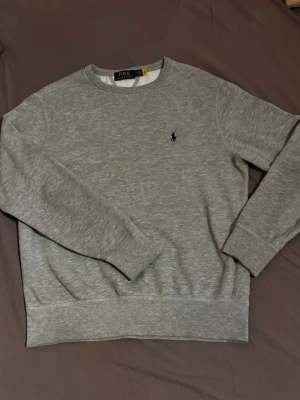 Grå sweatshirt från Polo Ralph Lauren - Klassisk grå sweatshirt från Polo Ralph Lauren med rund halsringning och diskret blå logga broderad på bröstet. Tröjan har ribbade muddar vid ärmslut och nederkant, perfekt för en avslappnad och stilren look.                                                                    Skicket skulle jag säga är 9/10 då den endast är använd några få gånger efter köp😄