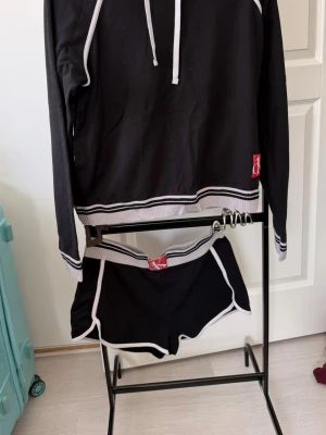 Svart loungewear set med vita detaljer - Snyggt svart loungewear set med hoodie och shorts. Setet har vita kontrastdetaljer längs kanterna och ribbade muddar. Hoodien har dragsko i halsen och båda plaggen har en liten röd logolapp. Perfekt för chill dagar hemma.