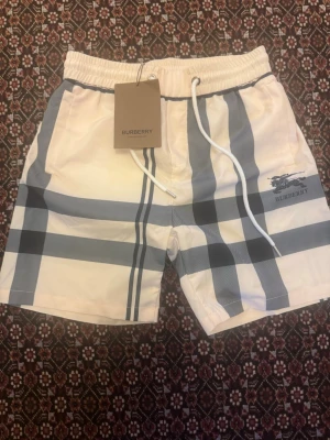 Rutig shorts med snörning Burberry - Säljer ett par snygga shorts från Burberry med klassiskt rutigt mönster i beige, svart och grått. Shortsen har elastisk midja med vit snörning och sidofickor. Perfekta för sommaren och ger en clean och trendig look.
