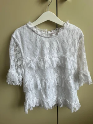 Vit spetsblus från Gina Tricot - Säljer en superfin vit spetsblus från Gina Tricot. Blusen har halvtransparent spets med blommönster, volangdetaljer och korta ärmar. Perfekt att styla med jeans eller kjol för en trendig look.