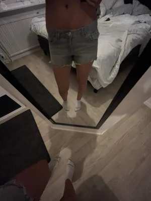 Grå jeansshorts med broderade fickor - Säljer ett par grå jeansshorts med rå kant och snygga broderade detaljer på bakfickorna. Shortsen har normal passform, bälteshällor och knäppning fram. Perfekta för sommaren och ger en avslappnad vibe.