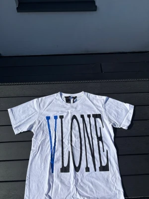 Vit VLONE t-shirt med tryck - Säljer en vit t-shirt från VLONE med stort svart och blått tryck framtill och en stor blå V med ormmotiv på ryggen. Klassisk passform och rund hals. Perfekt för dig som gillar streetwear och vill sticka ut med en unik design.