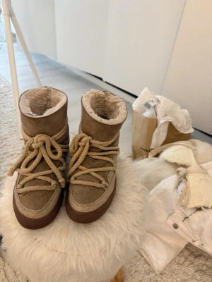 Beige Inuikiis - Säljer ett par beige moon boots med tjocka snören och mjukt fuskpälsfoder. Skorna har en högre modell och grov sula i brunt. Utsidan är i mockaliknande material och de är perfekta för kalla dagar.
