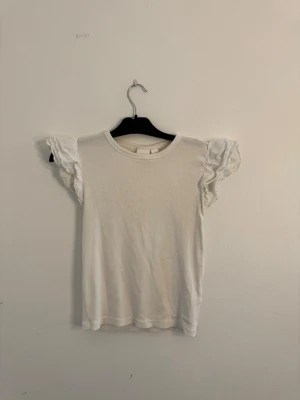 Vit topp med volangärmar - Söt vit topp med rund halsringning och korta volangärmar. Perfekt för dig som gillar en enkel men ändå feminin stil.