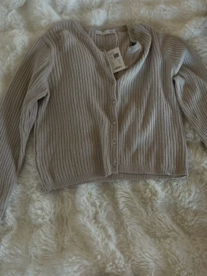 Beige ribbad kofta med knappar - Säljer en beige ribbad kofta med långa ärmar och v-ringning. Koftan har en croppad passform och stängs med små knappar framtill. Perfekt att slänga över en topp för en chill look.