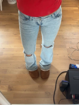 Ljusblå bootcut jeans med hål - Säljer ett par ljusblå bootcut jeans med slitningar över knäna och coola rosa sömmar på bakfickan. Jeansen har klassisk femficksdesign och är tillverkade i denim med en avslappnad passform. Perfekta för en trendig och avslappnad look. Säljer pga för små❣️